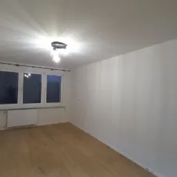 Pusty pok&oacute;j po remoncie: białe ściany, drewnopodobna podłoga, okno z grzejnikiem i nowoczesna lampa sufitowa.