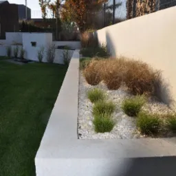 Nowoczesny ogród z podwyższonymi rabatami wyłożonymi białym żwirem i ozdobnymi trawami, ogrodzony metalową siatką i murem.