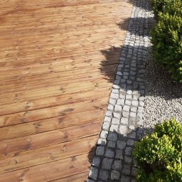 Ogrody z duszą Nie przesadzaj S.C - Widok z góry na drewniany taras z desek, graniczący z kostką brukową i żwirem, obok którego rosną zielone krzewy.