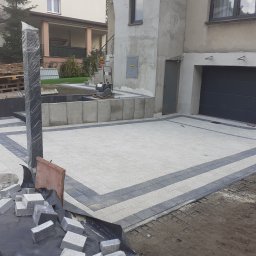 Ogrody z duszą Nie przesadzaj S.C - Ułożona kostka brukowa na podjeździe przed domem z garażem, widoczne materiały budowlane i narzędzia, słupek ogrodzeniowy owinięty folią zabezpieczającą.