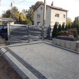 Ogrody z duszą Nie przesadzaj S.C - Nowo zamontowana, czarna, przesuwna brama ogrodzeniowa, jeszcze owinięta folią ochronną, na tle świeżo ułożonej kostki brukowej i częściowo zagospodarowanego terenu z tujami i paletami, widoczny...