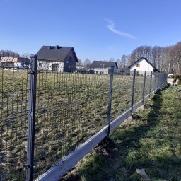 Ogrody z duszą Nie przesadzaj S.C - Nowo postawione, czarne ogrodzenie panelowe z betonową podmurówką na nierównym terenie, w tle domy jednorodzinne i pracownik z wiadrem.