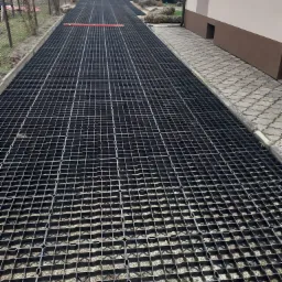 Ułożona, czarna, plastikowa kratka na podbudowie z kruszywa, tworząca utwardzoną powierzchnię wzdłuż budynku, z widocznymi pracownikami i sprzętem budowlanym w tle.