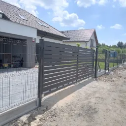 Nowoczesne ogrodzenie posesji z przesuwną bramą w kolorze antracytowym, widok z zewnątrz, z workami cementu w tle.