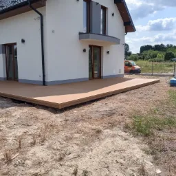 Nowoczesny taras z desek kompozytowych przylegający do białego domu z szarą podmurówką, widoczny pomarańczowy samochód dostawczy w tle, teren wokół tarasu jeszcze niezagospodarowany.