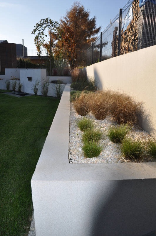 Nowoczesny ogród z podwyższonymi rabatami wyłożonymi białym żwirem i ozdobnymi trawami, ogrodzony metalową siatką i murem.