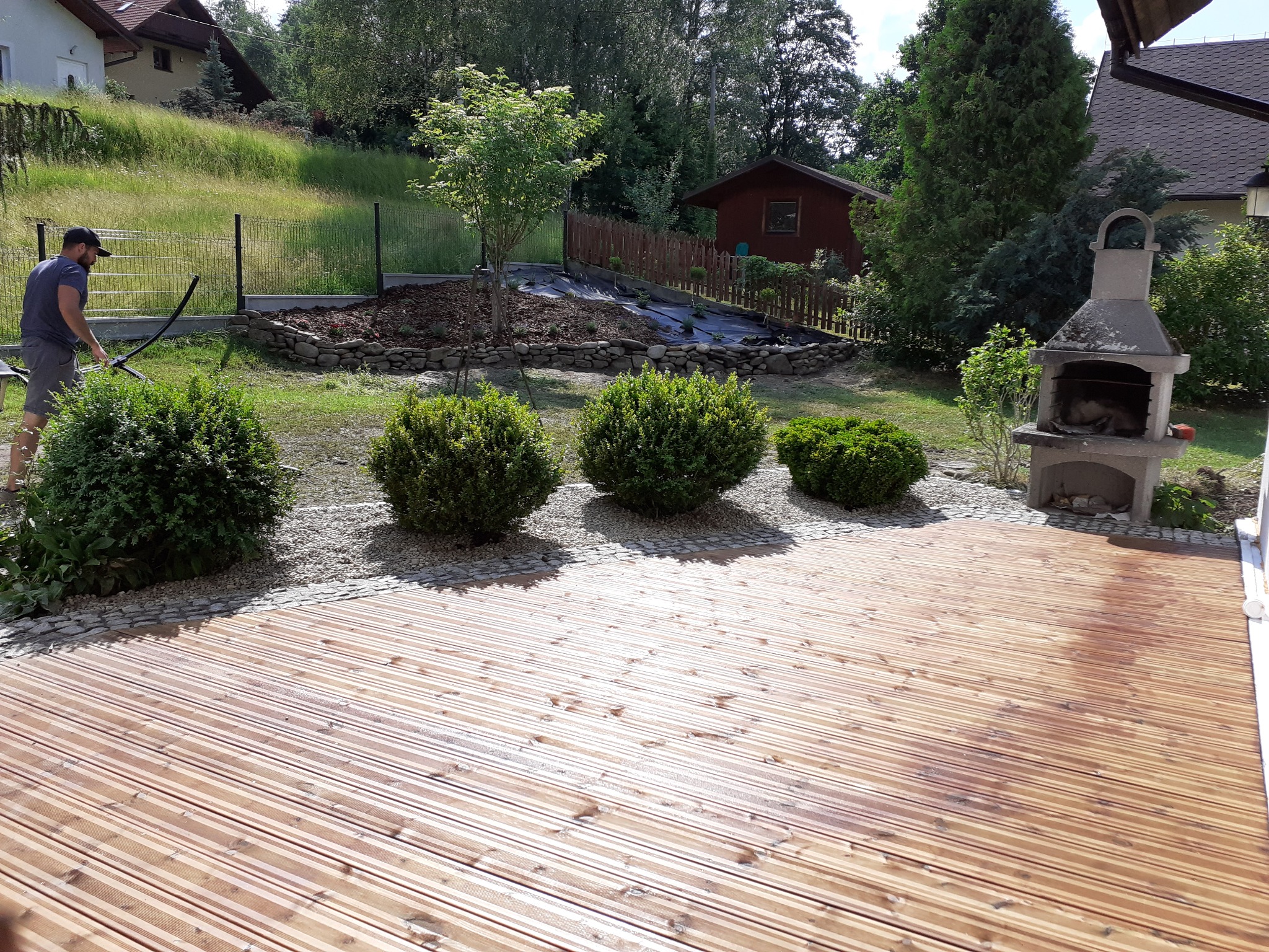 Nowo wybudowany drewniany taras z czterema kulistymi krzewami ozdobnymi, kamienną obwódką i grillem betonowym w tle, w oddali widoczny mężczyzna przy ogrodzeniu z siatki.