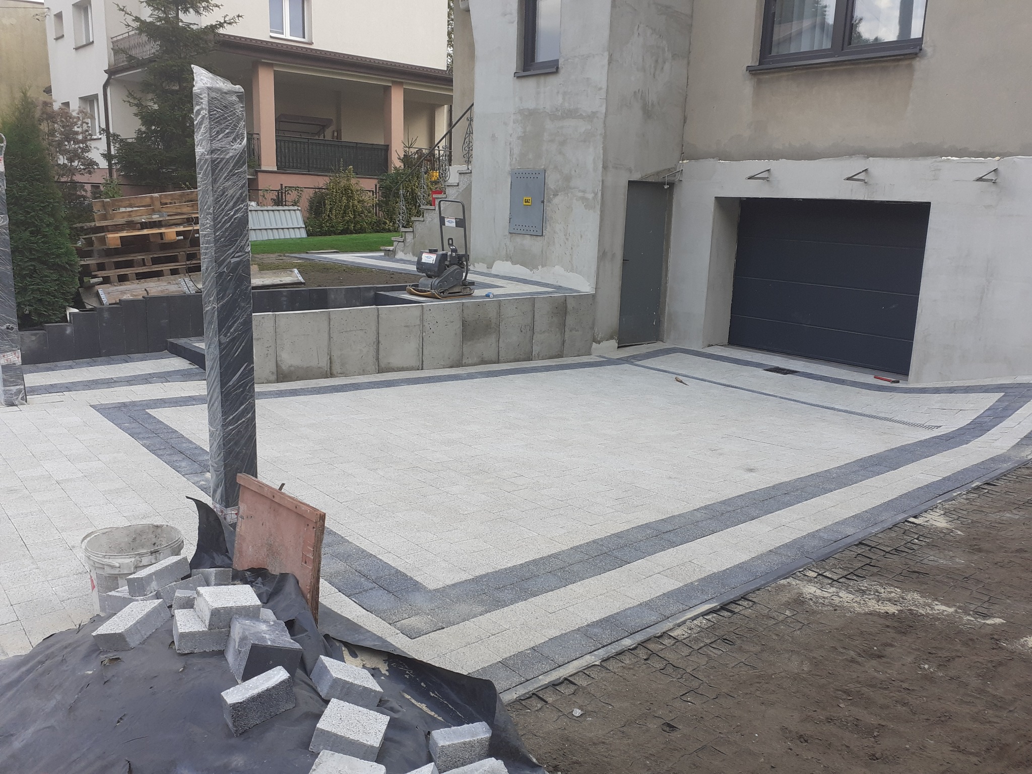 Ułożona kostka brukowa na podjeździe przed domem z garażem, widoczne materiały budowlane i narzędzia, słupek ogrodzeniowy owinięty folią zabezpieczającą.