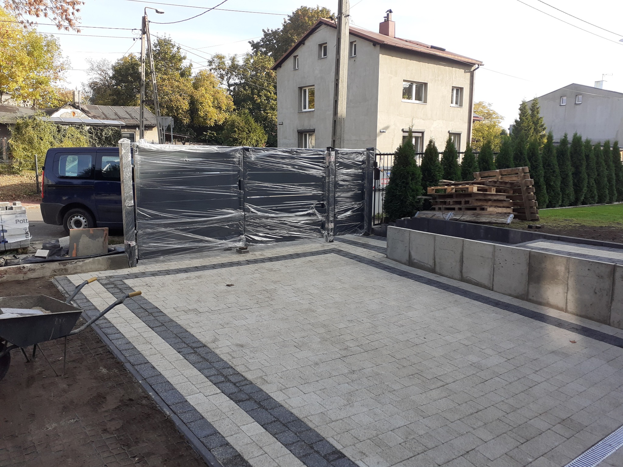 Nowo zamontowana, czarna, przesuwna brama ogrodzeniowa, jeszcze owinięta folią ochronną, na tle świeżo ułożonej kostki brukowej i częściowo zagospodarowanego terenu z tujami i paletami, widoczny...