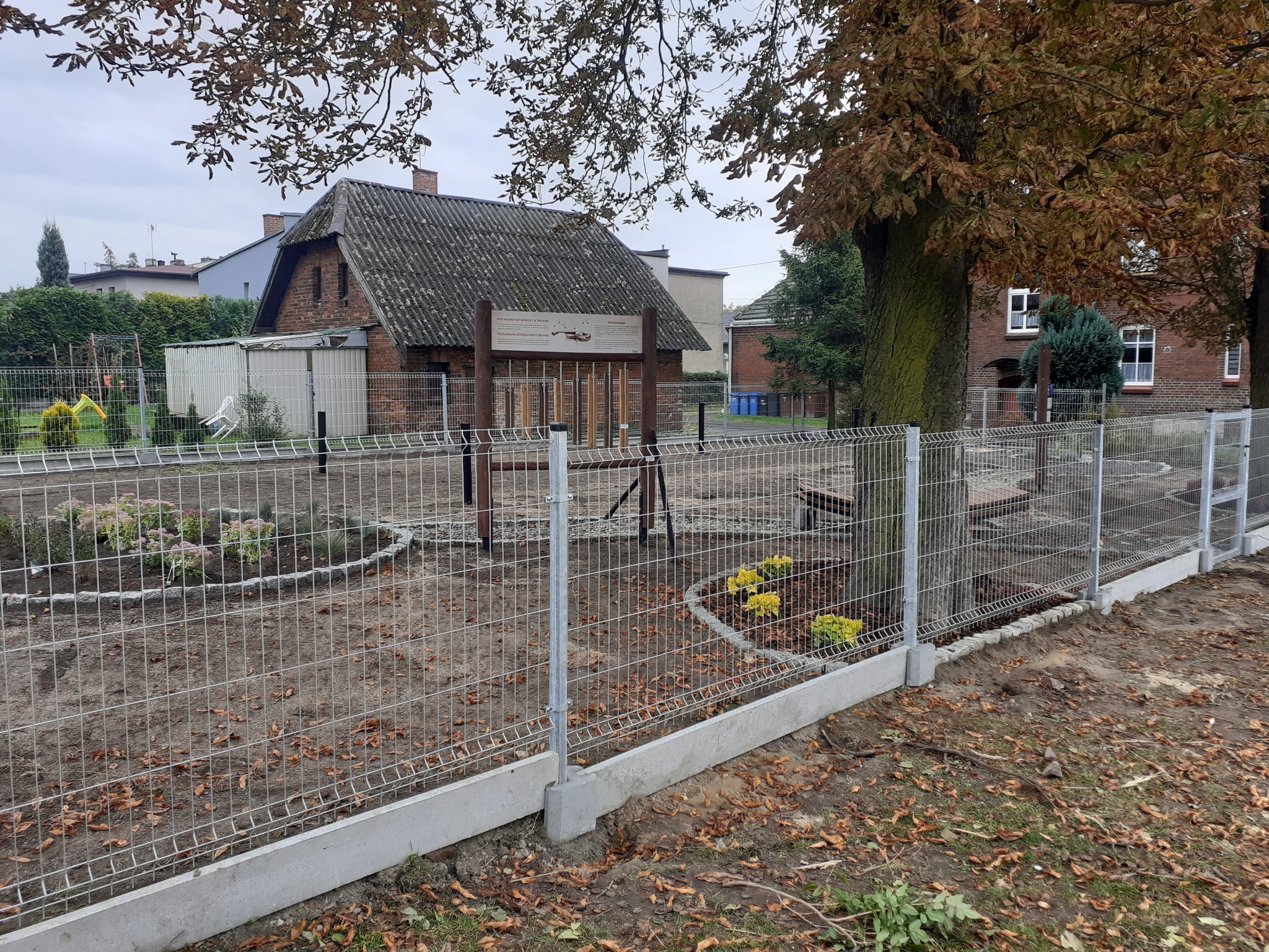 Nowo zamontowane, metalowe ogrodzenie panelowe z podmurówką betonową, otaczające teren z rabatami kwiatowymi i drzewami, w tle budynki mieszkalne.