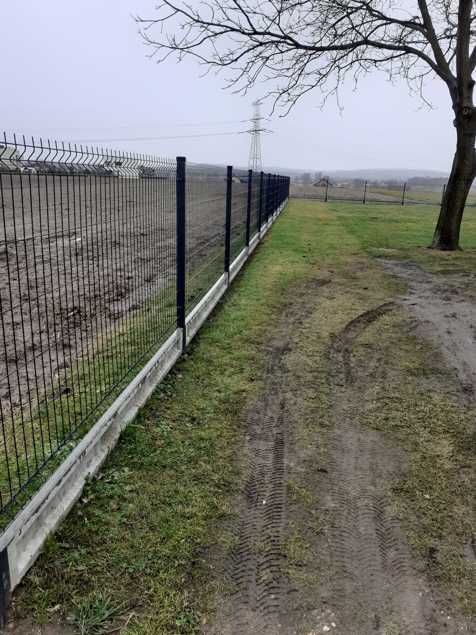 Nowoczesne ogrodzenie panelowe z podmurówką betonową, widoczne ślady opon na trawie i drzewo bez liści w tle.