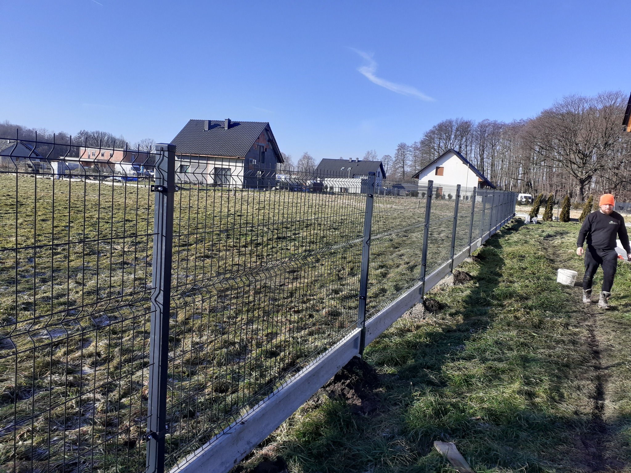 Nowo postawione, czarne ogrodzenie panelowe z betonową podmurówką na nierównym terenie, w tle domy jednorodzinne i pracownik z wiadrem.