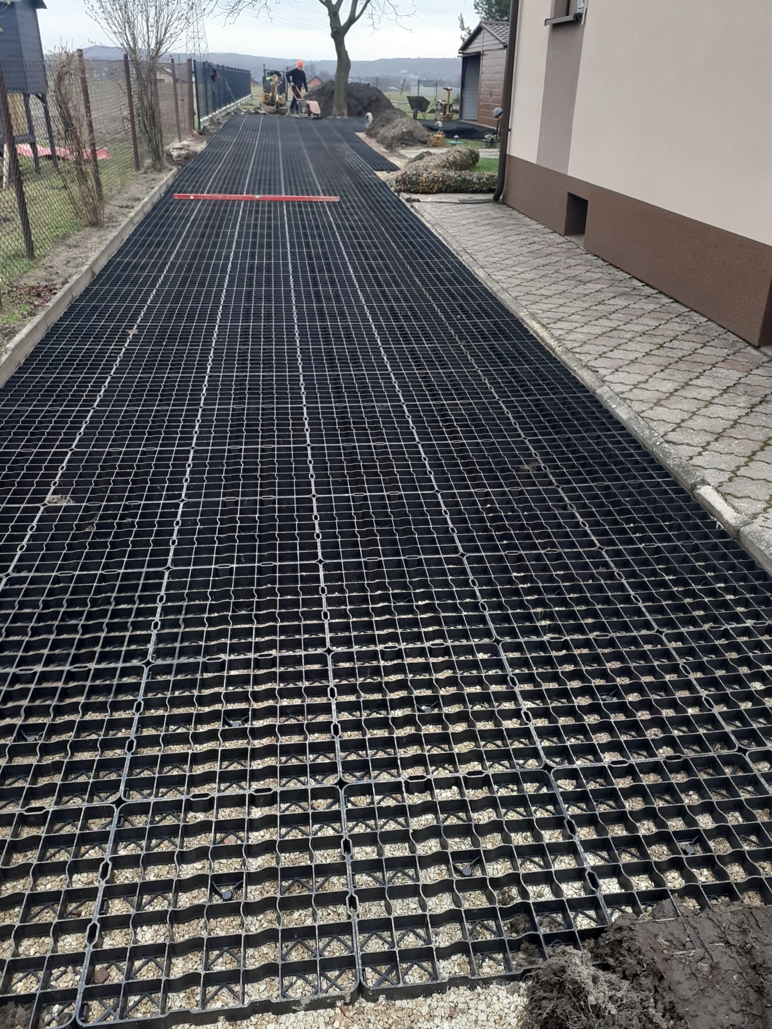 Ułożona, czarna, plastikowa kratka na podbudowie z kruszywa, tworząca utwardzoną powierzchnię wzdłuż budynku, z widocznymi pracownikami i sprzętem budowlanym w tle.