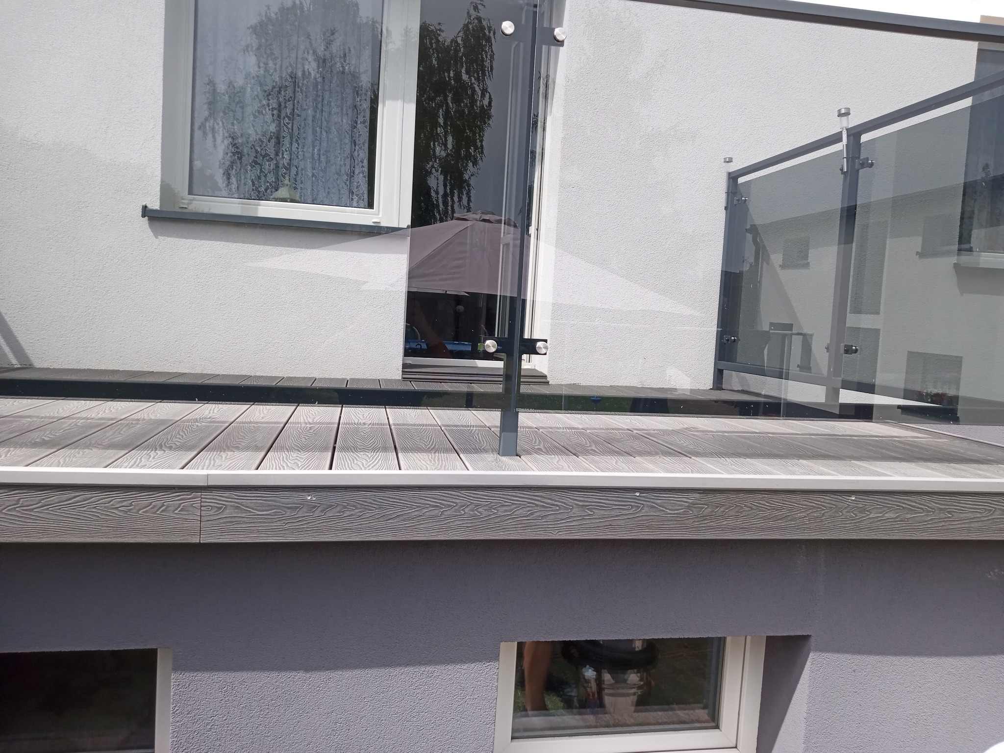 Nowoczesny taras z szarą deską kompozytową i szklaną balustradą, widok z dołu na elewację budynku z oknami.