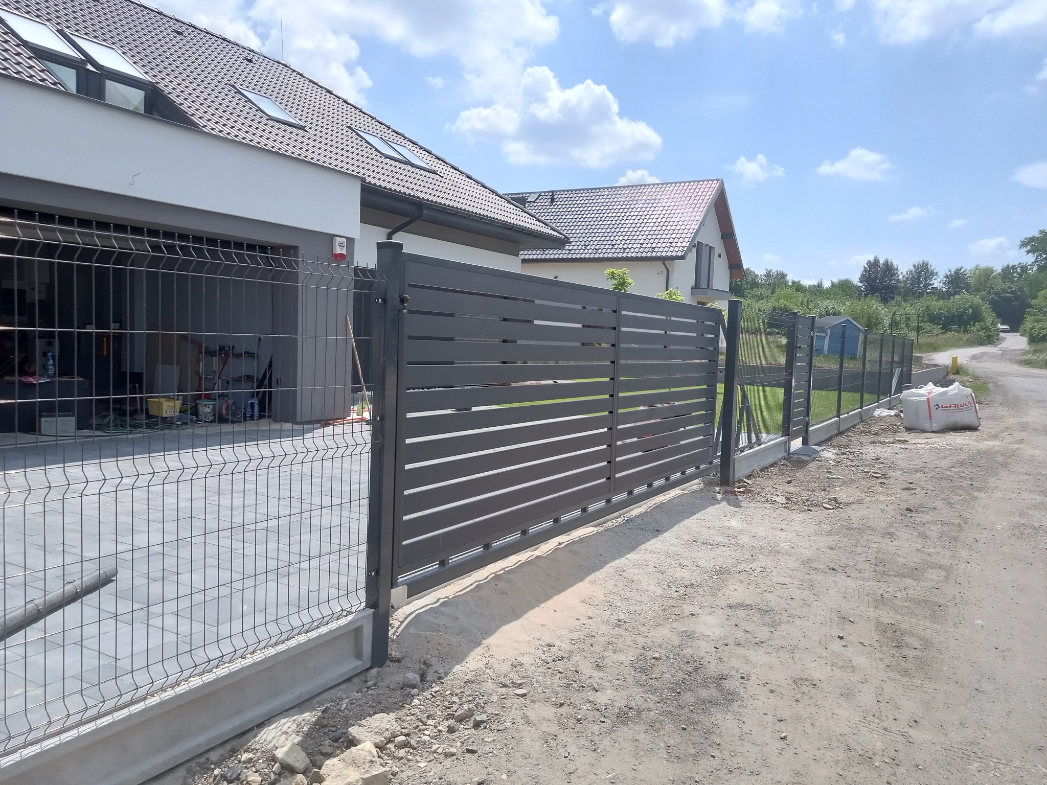 Nowoczesne ogrodzenie posesji z przesuwną bramą w kolorze antracytowym, widok z zewnątrz, z workami cementu w tle.