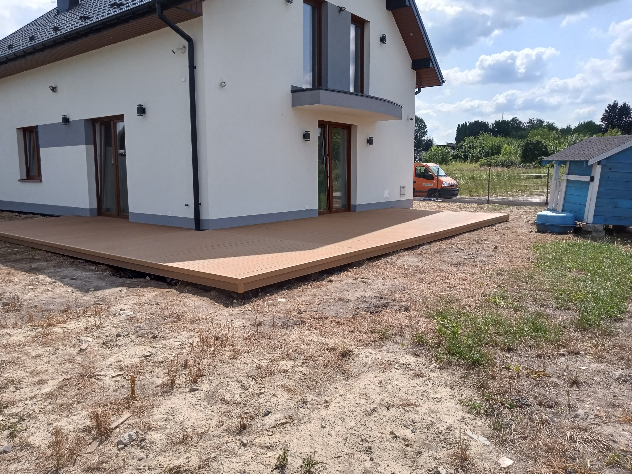 Nowoczesny taras z desek kompozytowych przylegający do białego domu z szarą podmurówką, widoczny pomarańczowy samochód dostawczy w tle, teren wokół tarasu jeszcze niezagospodarowany.