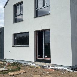 Building-fach Filip Chorążewicz - Surowy stan domu jednorodzinnego z czarnymi oknami i szarym dachem, po ociepleniu elewacji. Widoczne deski i gruz na ziemi.