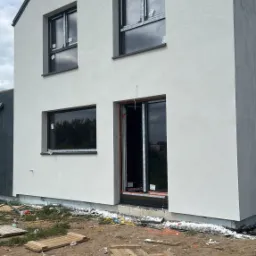 Surowy stan domu jednorodzinnego z czarnymi oknami i szarym dachem, po ociepleniu elewacji. Widoczne deski i gruz na ziemi.