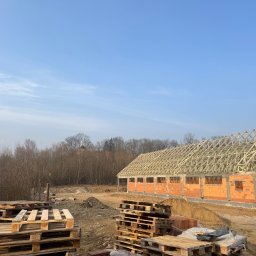 Building-fach Filip Chorążewicz - Budowa domu z czerwonej cegły: ściany wzniesione, drewniana konstrukcja dachu w trakcie montażu. Na pierwszym planie ułożone palety. W tle drzewa i błękitne niebo.