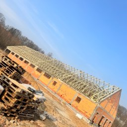 Building-fach Filip Chorążewicz - Wznoszenie budynku z cegły: ściany z otworami okiennymi i drewniana konstrukcja dachu pod błękitnym niebem. Na pierwszym planie palety budowlane.