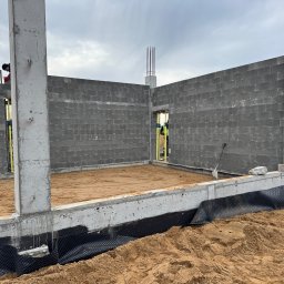 Building-fach Filip Chorążewicz - Budowa domu: ściany z szarego betonu komórkowego, zbrojone betonowe słupy, fundament z izolacją przeciwwilgociową i piaskiem wewnątrz. Widoczny pracownik w tle.