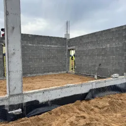 Budowa domu: ściany z szarego betonu komórkowego, zbrojone betonowe słupy, fundament z izolacją przeciwwilgociową i piaskiem wewnątrz. Widoczny pracownik w tle.