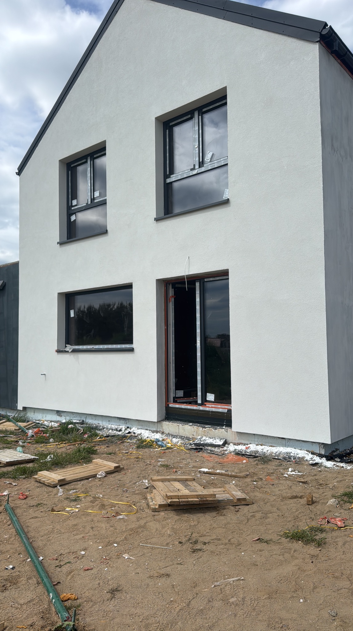 Surowy stan domu jednorodzinnego z czarnymi oknami i szarym dachem, po ociepleniu elewacji. Widoczne deski i gruz na ziemi.