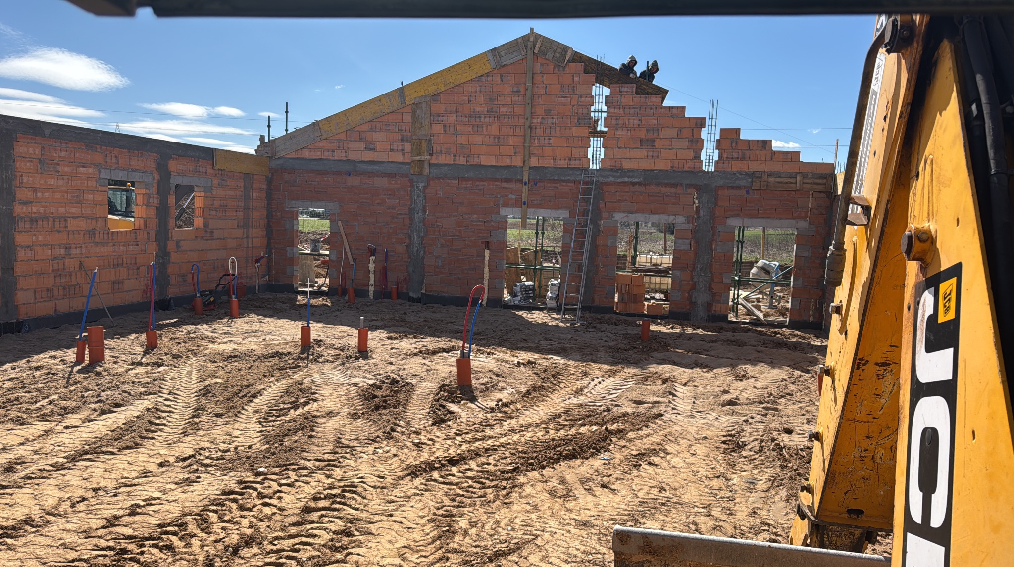 Budowa domu z czerwonej cegły, z widocznymi elementami konstrukcyjnymi i pracownikami na dachu. Na pierwszym planie ziemia z wyraźnymi śladami opon koparki.