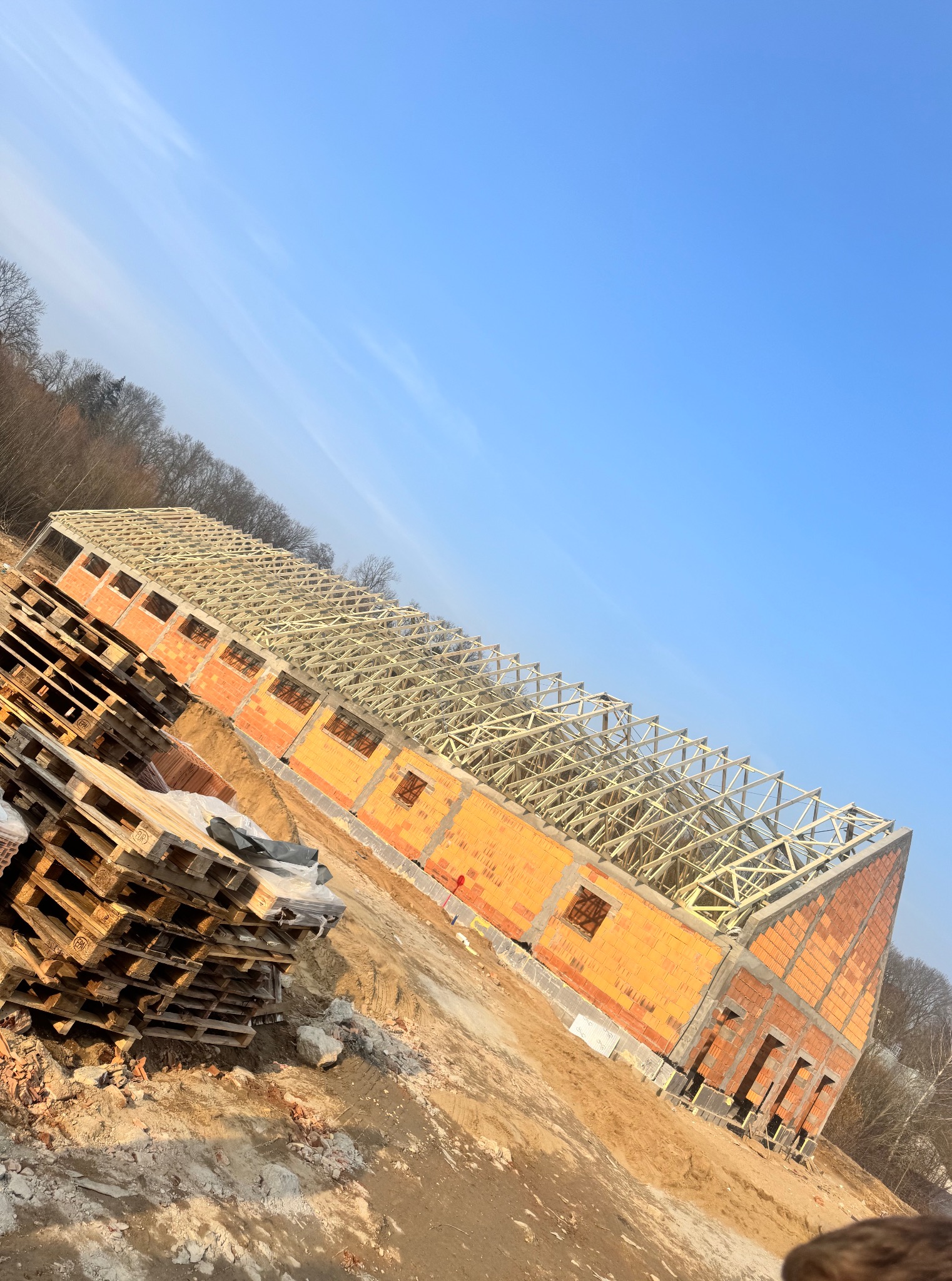 Wznoszenie budynku z cegły: ściany z otworami okiennymi i drewniana konstrukcja dachu pod błękitnym niebem. Na pierwszym planie palety budowlane.