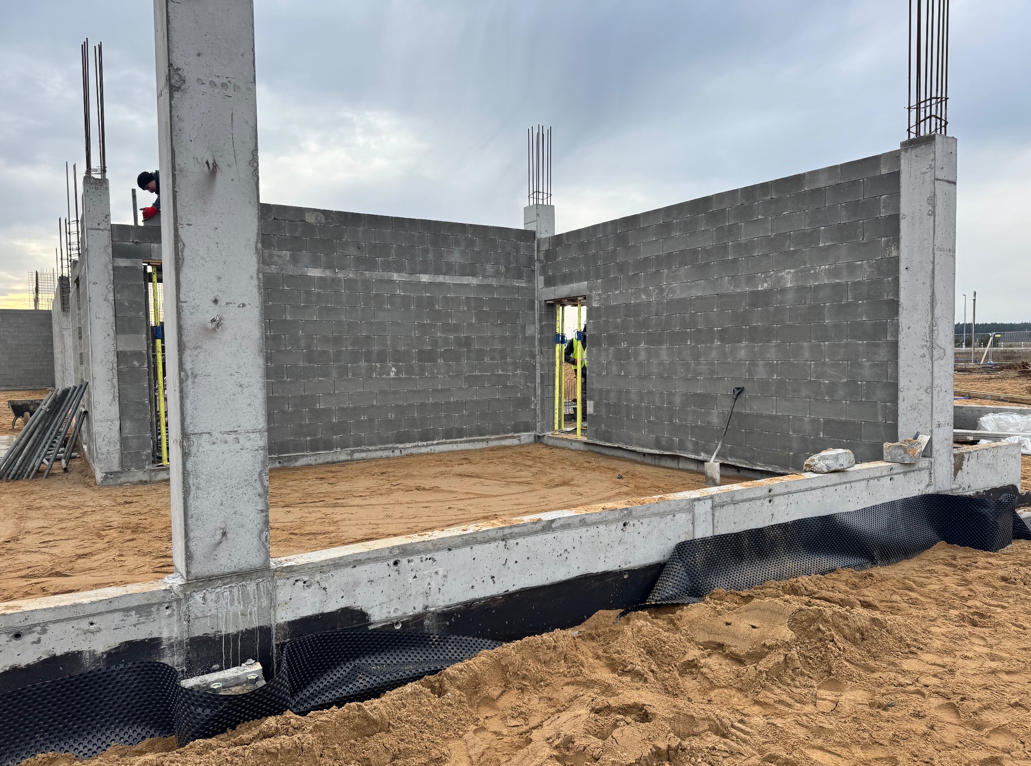 Budowa domu: ściany z szarego betonu komórkowego, zbrojone betonowe słupy, fundament z izolacją przeciwwilgociową i piaskiem wewnątrz. Widoczny pracownik w tle.