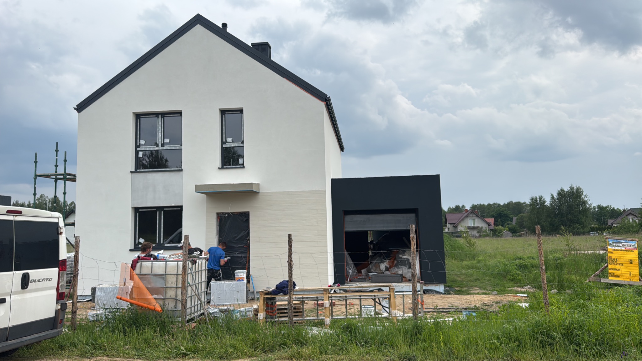 Dwukondygnacyjny dom z garażem w trakcie ocieplania elewacji. Widoczni pracownicy, materiały budowlane i samochód dostawczy. Część elewacji jest już ocieplona.