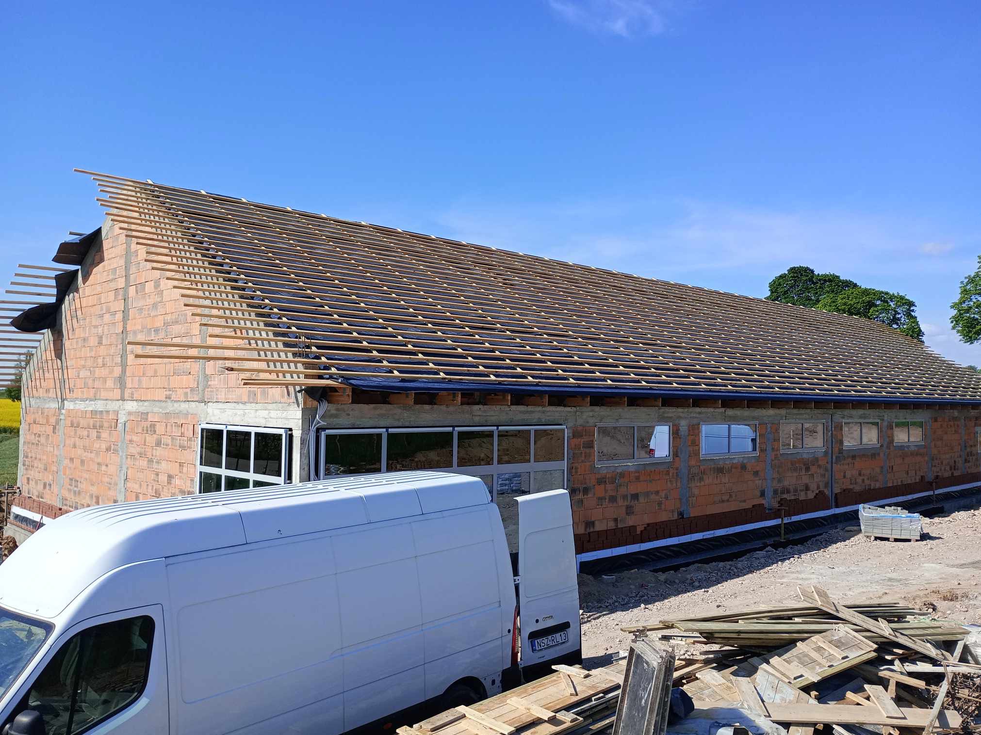Budynek w trakcie budowy z czerwonej cegły, widoczne okna i drewniana konstrukcja dachu. Na pierwszym planie biały bus dostawczy i materiały budowlane.