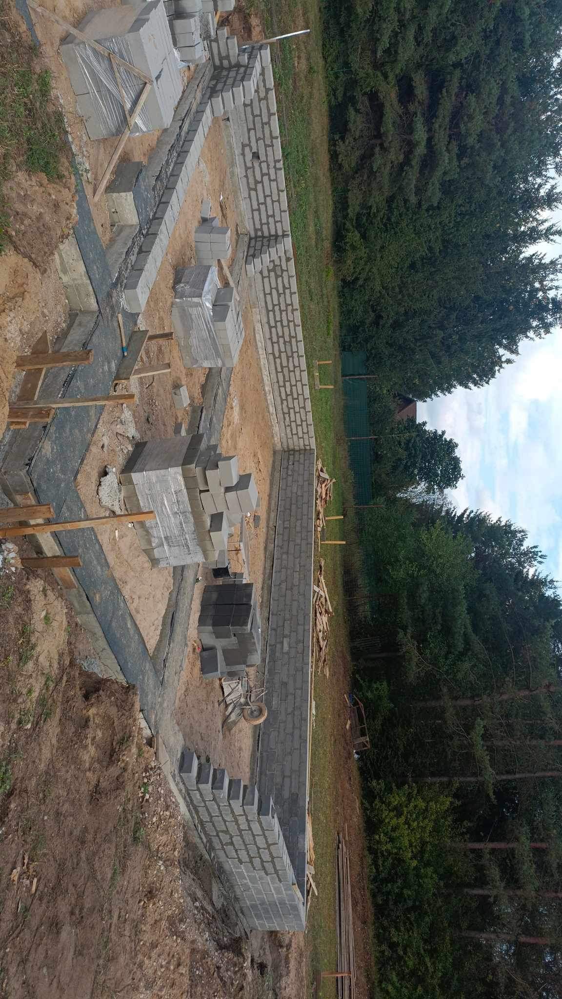 Budowa fundamentów z bloczków betonowych na leśnej działce. Częściowo wymurowane ściany fundamentowe, obok paleta z bloczkami. Widok z góry.