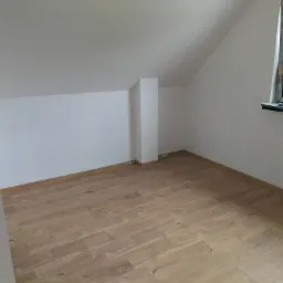 Poddasze po remoncie: świeżo pomalowane białe ściany i ułożona jasna podłoga panelowa, widoczne okno z widokiem na zieleń.