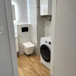 Wykończona łazienka w Lubinie z drewnopodobnymi panelami podłogowymi, wiszącą toaletą, pralką i marmurowymi płytkami ściennymi z widocznym bojlerem.