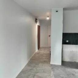 Wnętrze mieszkania po remoncie, świeżo pomalowane ściany w odcieniach szarości, ułożona szara podłoga z płytek, widoczne drzwi wewnętrzne i fragment przygotowywanej kuchni z czarnym blatem.