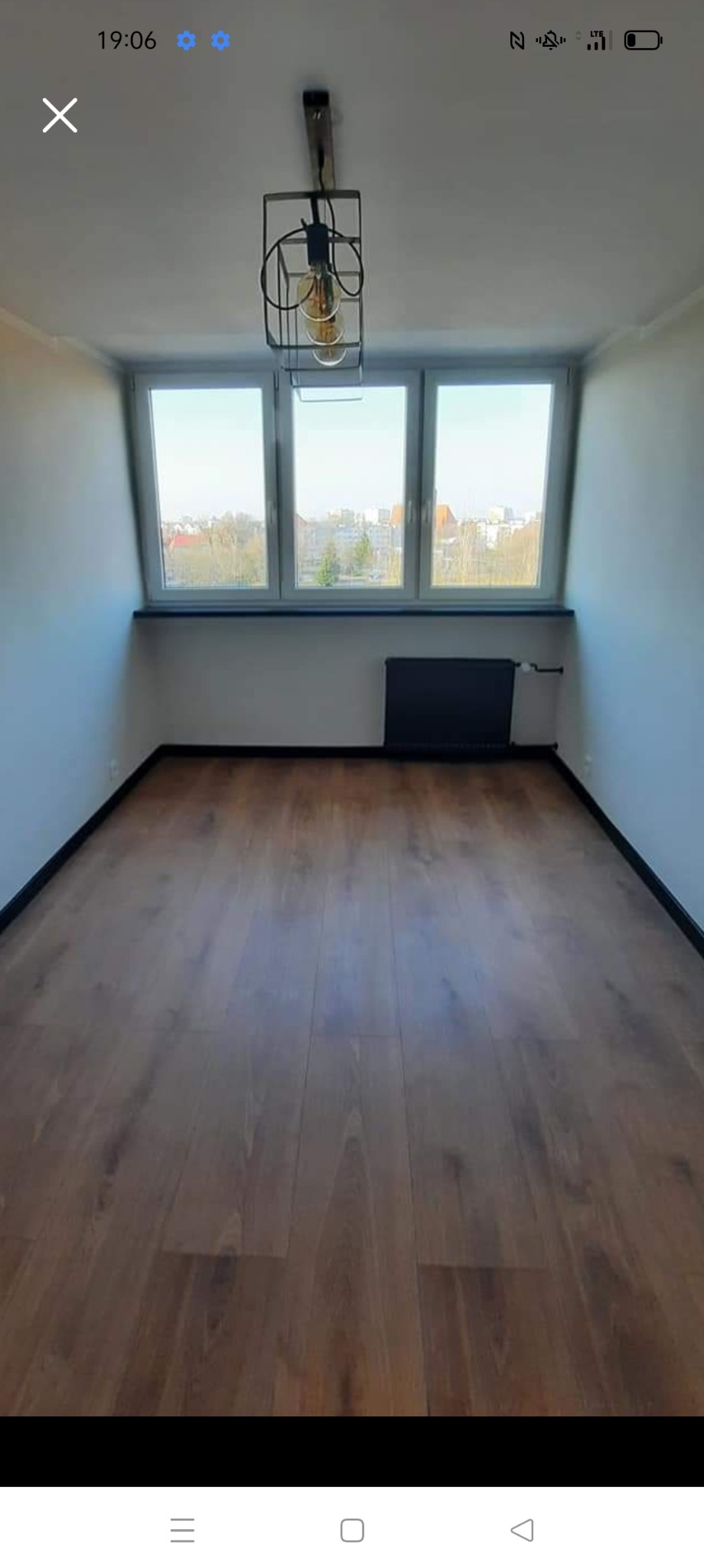 Pokój po remoncie z nową podłogą z paneli w odcieniach brązu, widok na okno z widokiem na miasto, nowoczesna lampa wisząca w stylu industrialnym i grzejnik w kolorze czarnym.