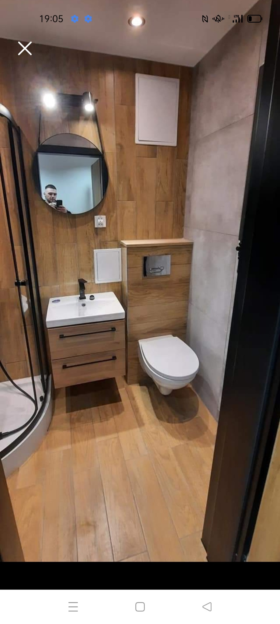 Nowoczesna łazienka z drewnopodobnymi płytkami na ścianach i podłodze, wisząca toaleta, szafka z umywalką i okrągłe lustro z odbijającym się fotografem.