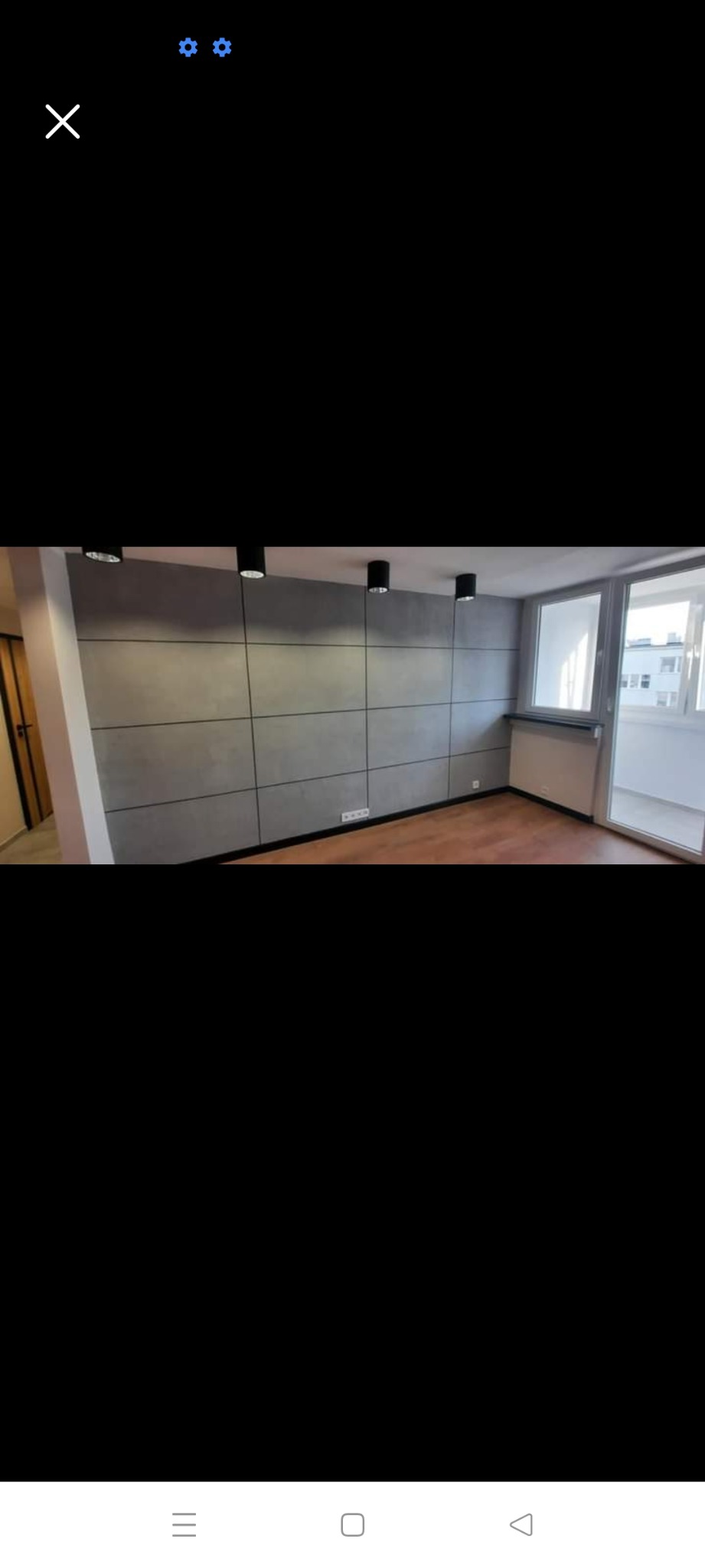 Pokój z panelami ściennymi imitującymi beton, oświetlenie punktowe na suficie, parkiet na podłodze, okno balkonowe z widokiem na zewnątrz.