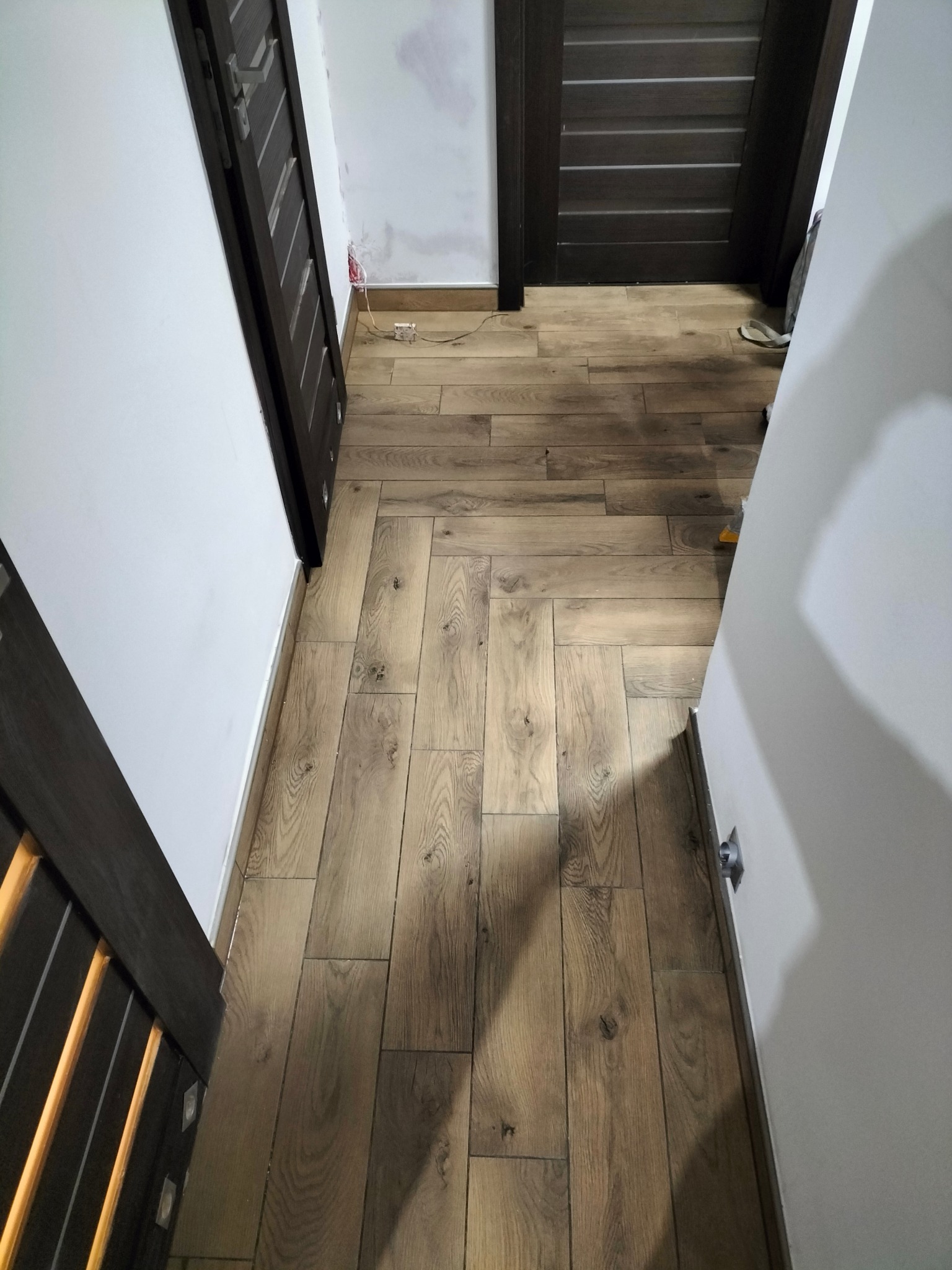 Ułożone panele podłogowe w korytarzu, widoczne fragmenty dwóch ciemnych drzwi, ściany częściowo przygotowane do malowania.