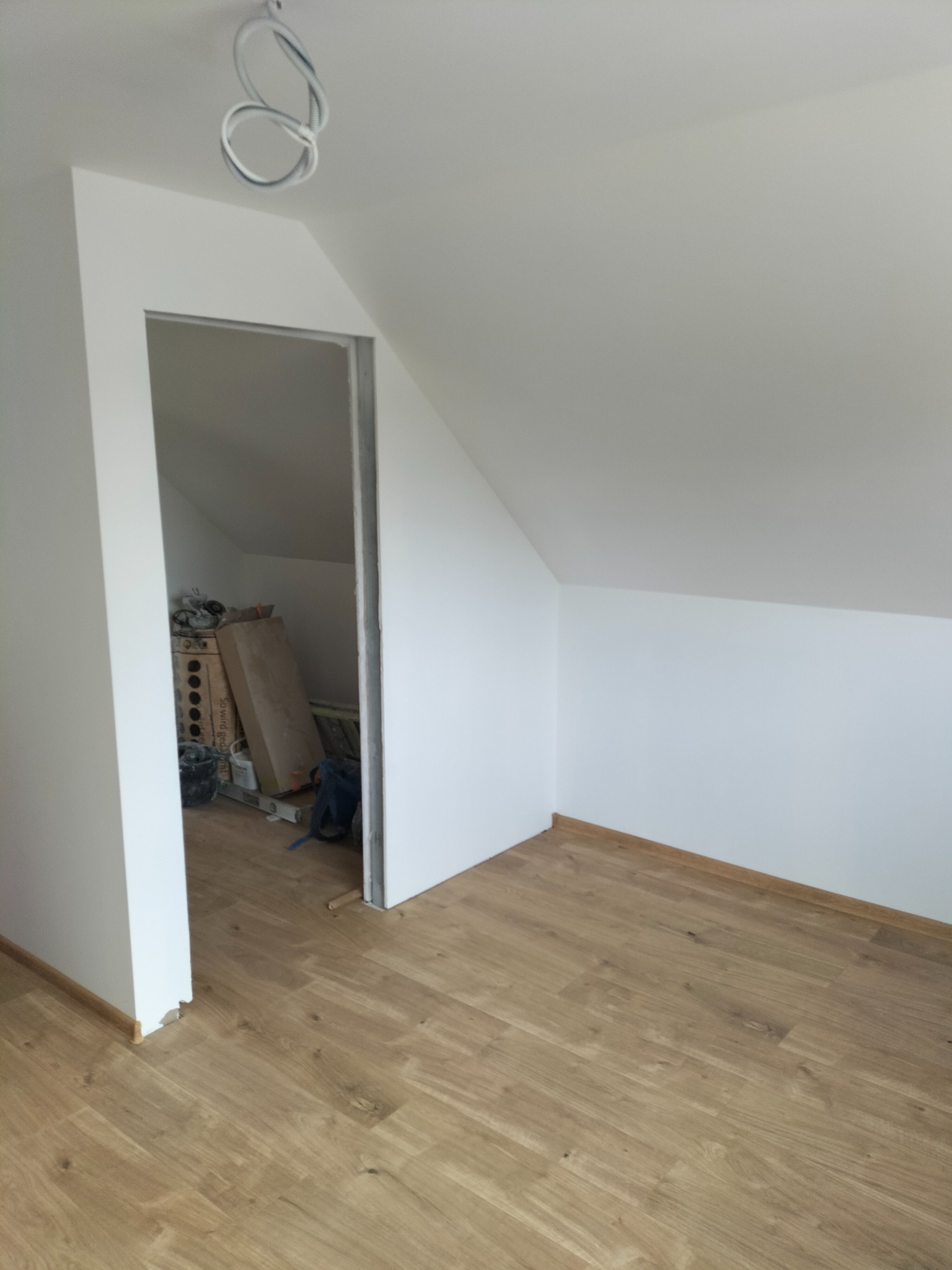 Poddasze po remoncie: świeżo ułożone panele podłogowe w jasnym odcieniu drewna, białe ściany i sufit, widoczne kable oświetleniowe oraz otwór do schowka z narzędziami i materiałami budowlanymi...