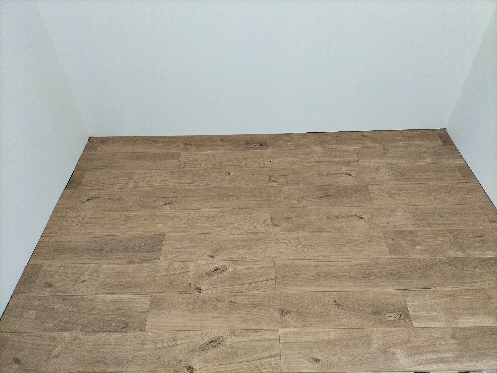 Jasnobrązowe panele podłogowe ułożone w rogu pomieszczenia z białymi ścianami, widoczne charakterystyczne sęki i wzory drewna.