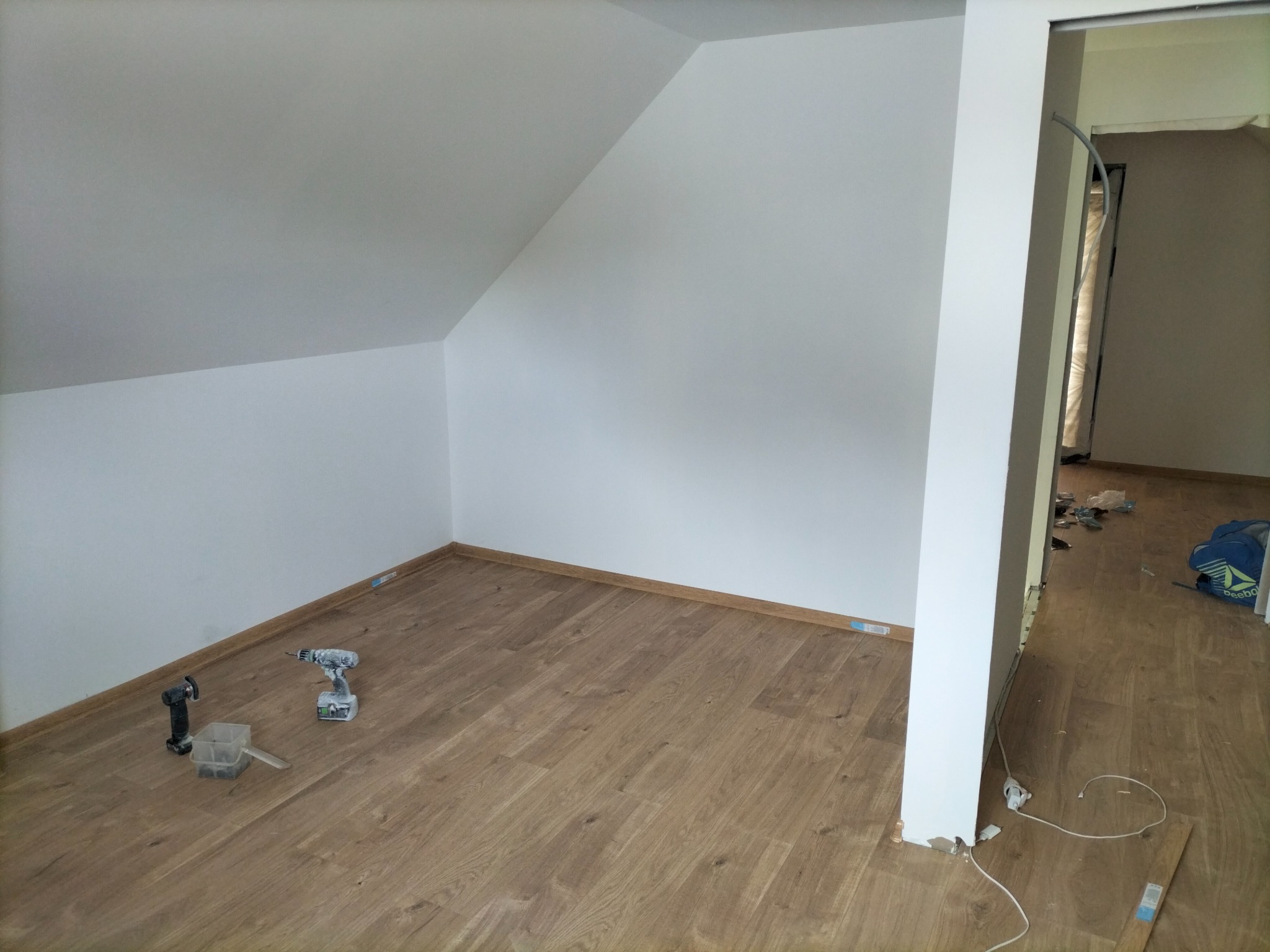 Poddasze z ułożonymi panelami podłogowymi, widoczne narzędzia: wiertarka i oscylacyjne narzędzie wielofunkcyjne, listwy przypodłogowe zamontowane, ściany pomalowane na biało, widoczny fragment...