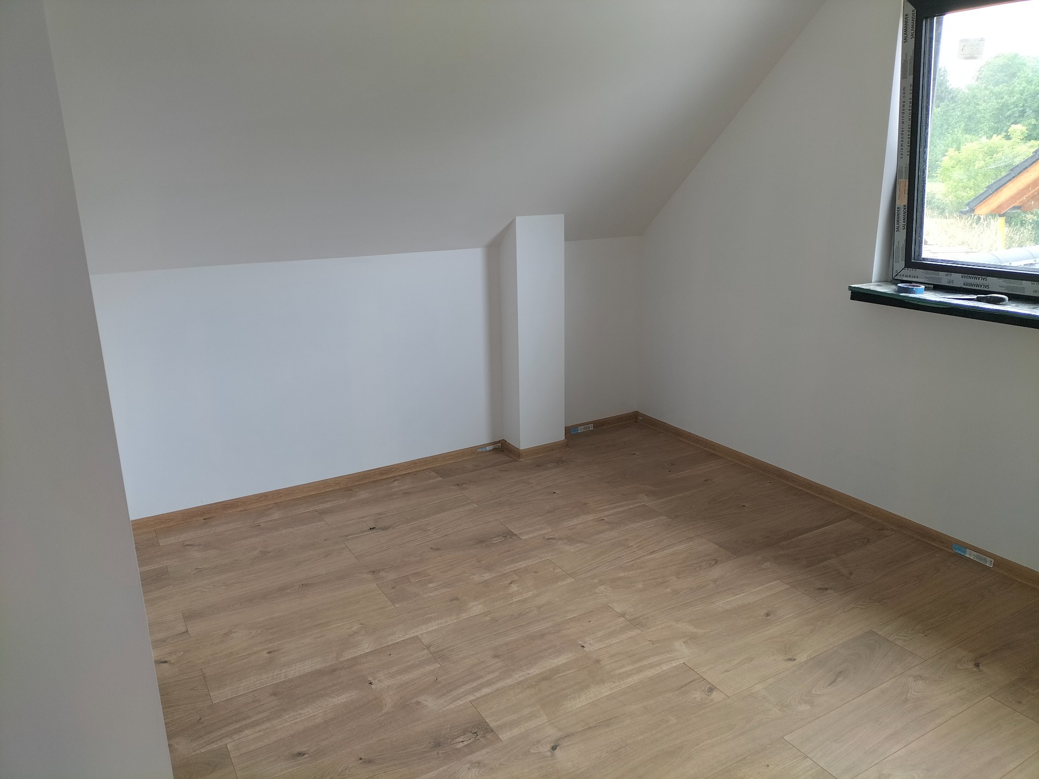 Poddasze po remoncie: świeżo pomalowane białe ściany i ułożona jasna podłoga panelowa, widoczne okno z widokiem na zieleń.