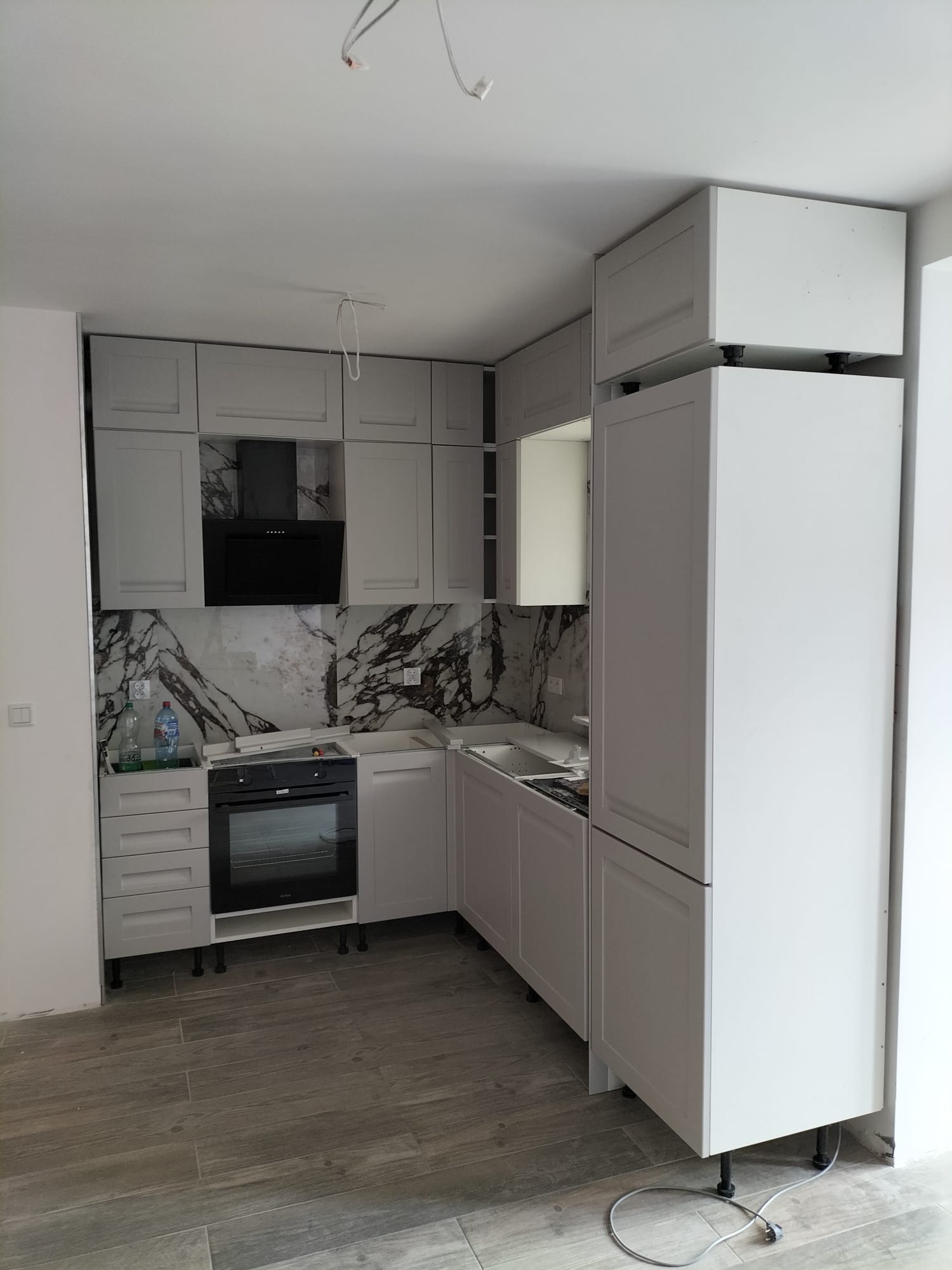 Montaż szarych szafek kuchennych z czarnym okapem i marmurowym panelem ściennym, widoczne niedokończone podłączenia elektryczne i butelki z napojami na blacie.