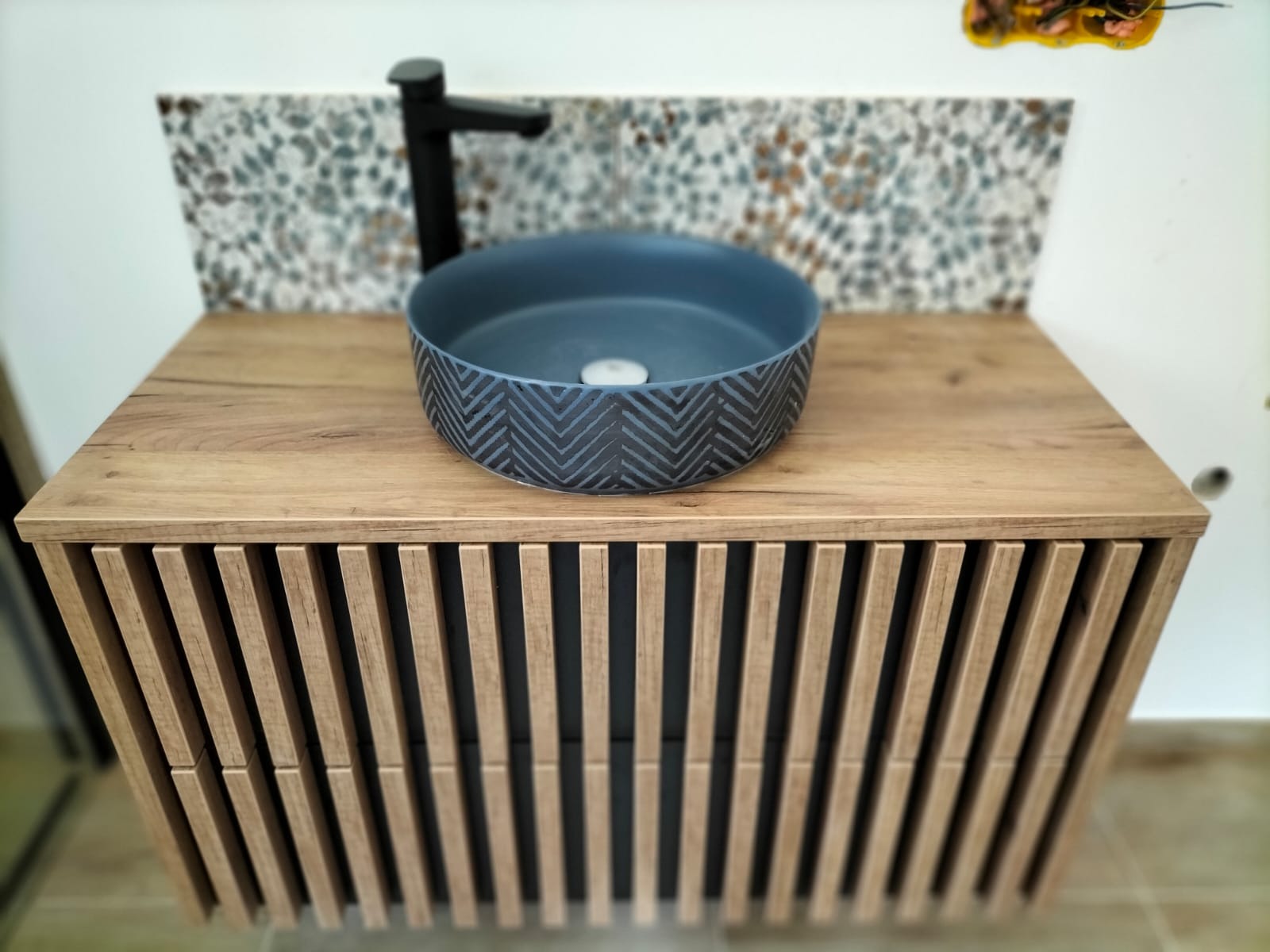 Nowoczesna łazienka z umywalką w kształcie misy z geometrycznym wzorem, zamontowaną na szafce z drewnianymi lamelami i czarną armaturą na tle mozaiki.