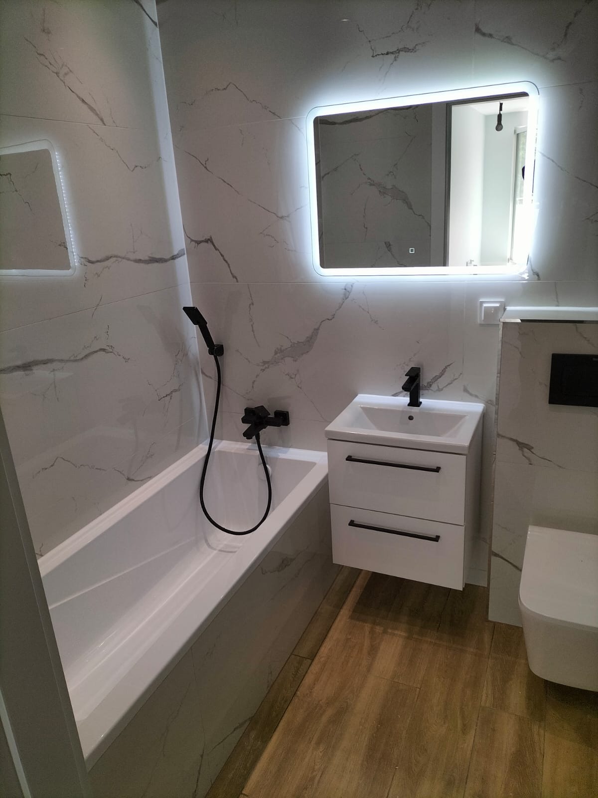 Wykończona łazienka z wanną, umywalką z szafką i toaletą, utrzymana w bieli i szarości z akcentami czerni, podświetlane lustro nad umywalką.