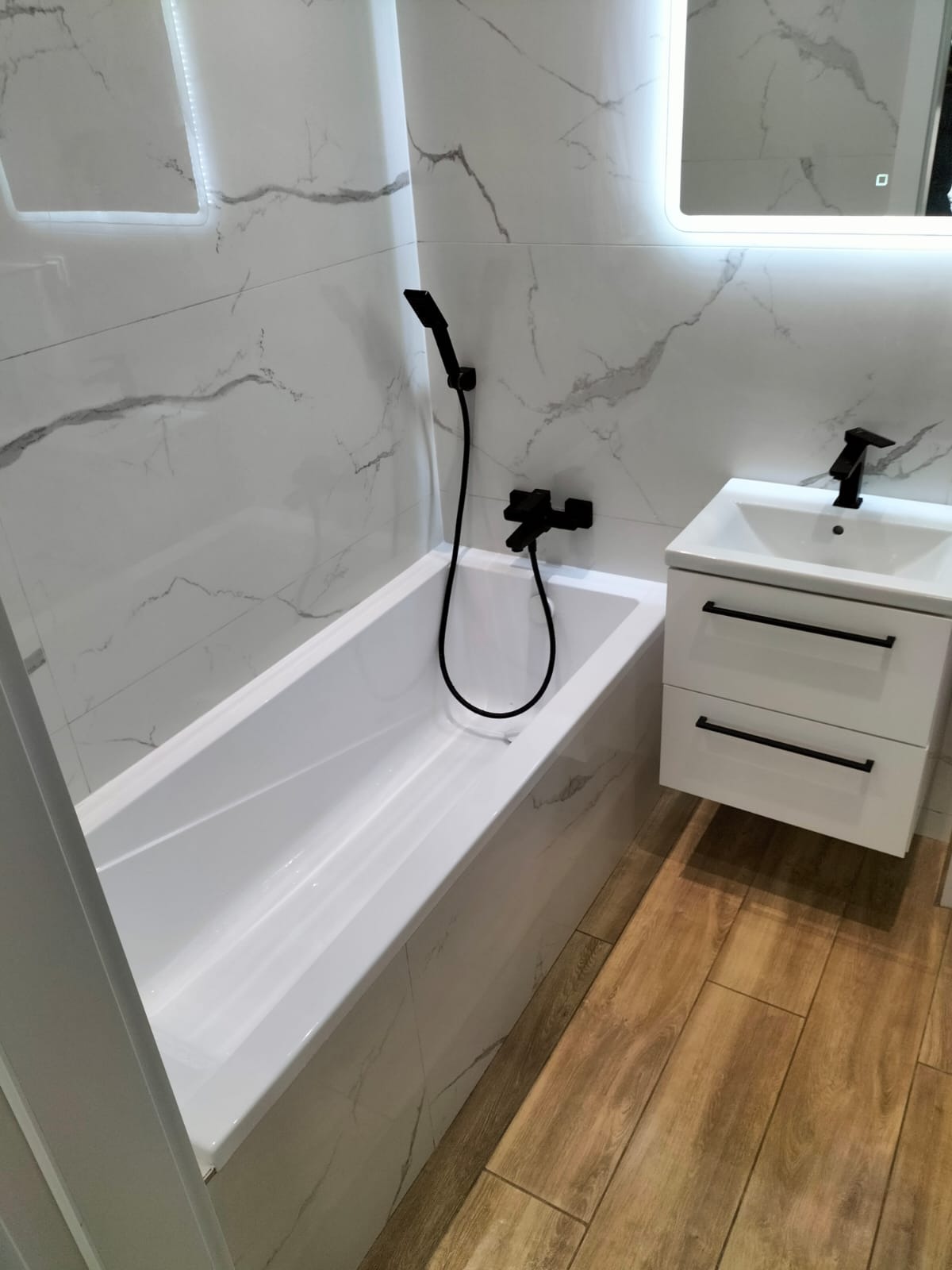 Nowoczesna łazienka z białą wanną, umywalką z szafką i czarną armaturą na tle płytek imitujących marmur i drewnopodobnej podłogi.