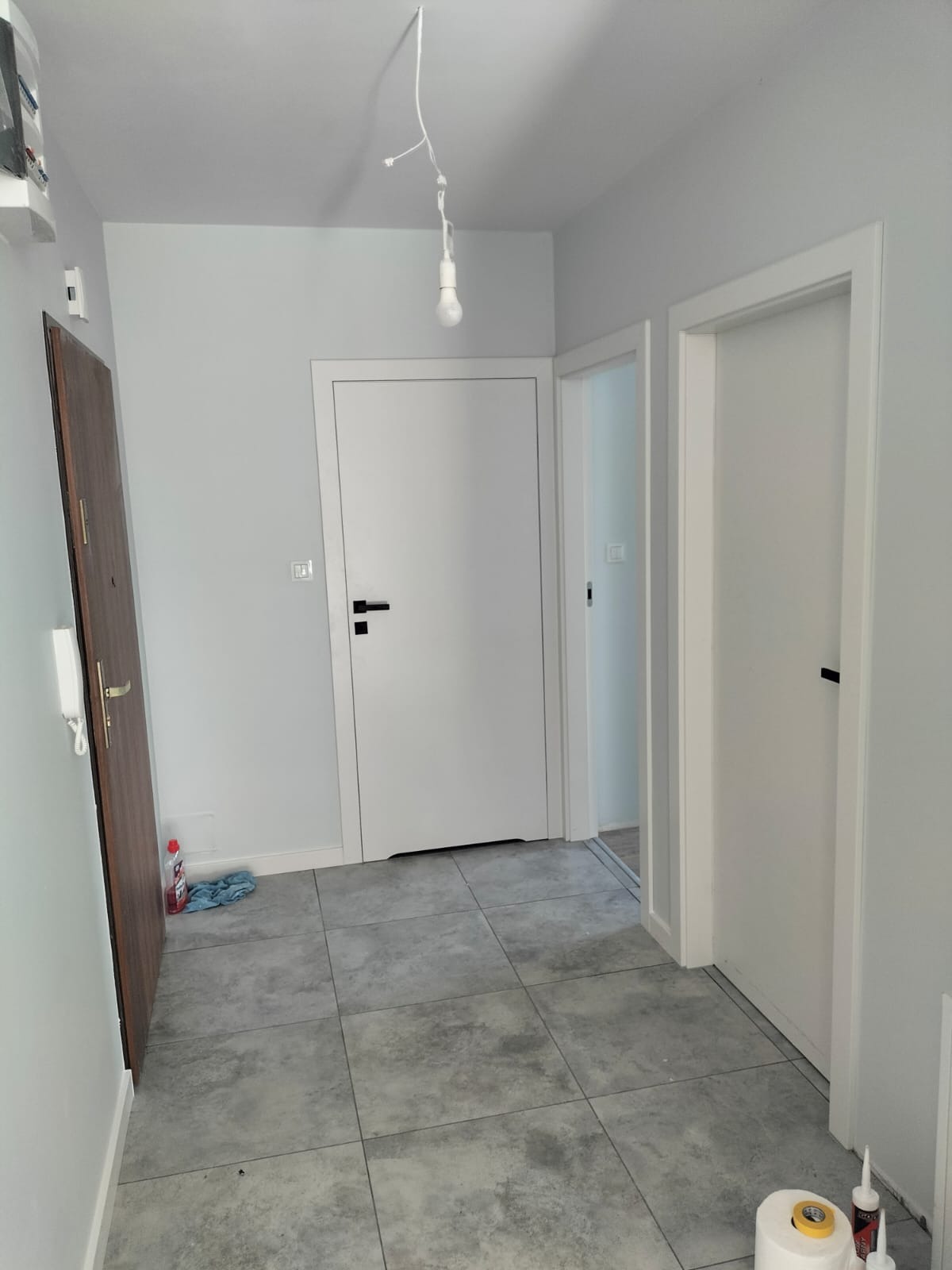 Korytarz po remoncie z szarymi płytkami podłogowymi, trzema parami drzwi w kolorze białym z czarnymi klamkami, gołą żarówką zwisającą z sufitu i butelką płynu do czyszczenia pod ścianą.
