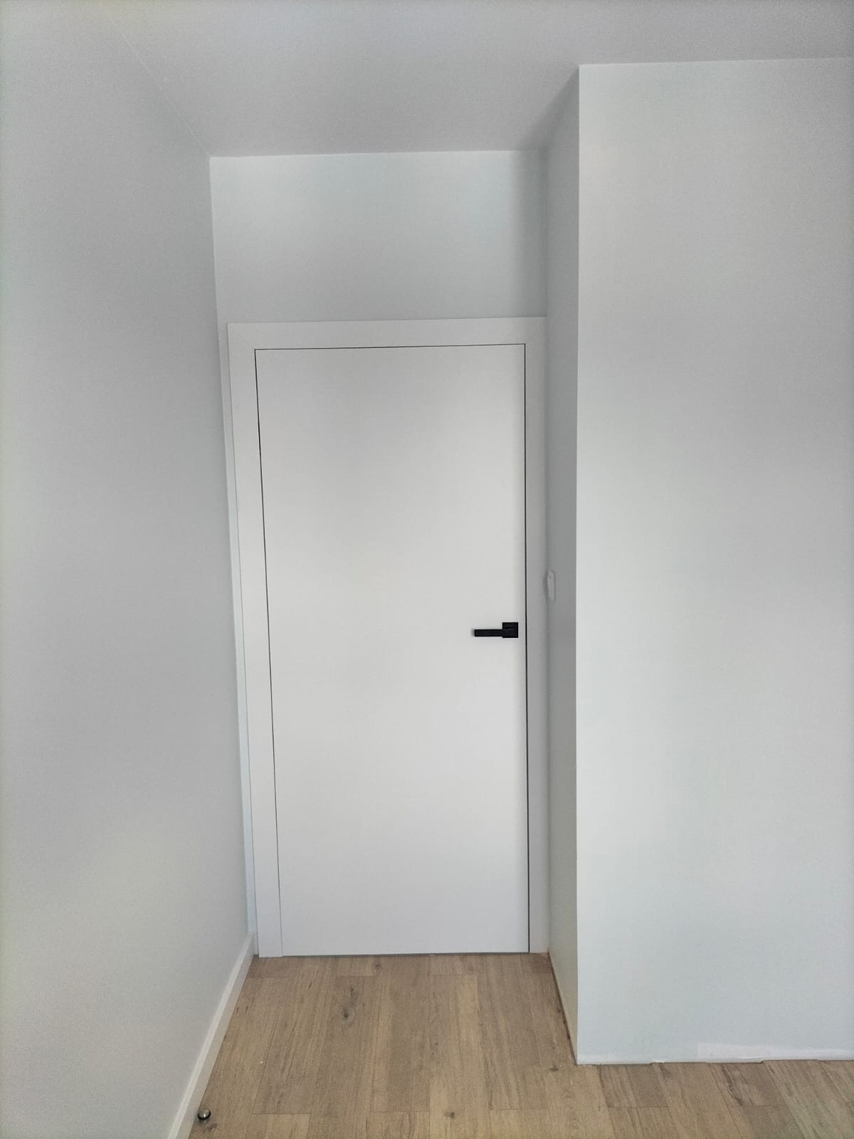 Minimalistyczne wnętrze z białą ścianą, drzwiami z czarną klamką i panelami podłogowymi w odcieniach drewna.